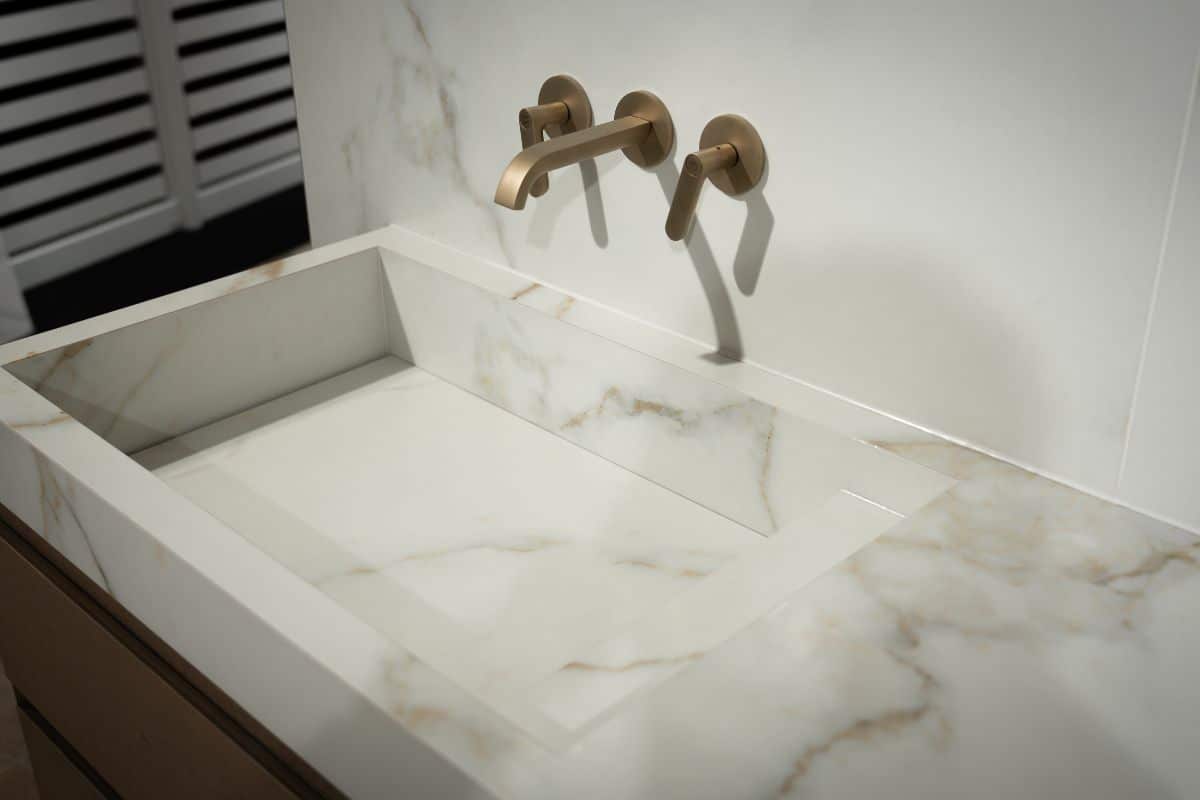 Waschtisch-Idee aus der Kr&uuml;ger Hannover Manufaktur: ma&szlig;gefertigter Waschtisch mit integriertem Waschbecken, alles gefertigt aus hellen Fliesen in eleganter Marmoroptik, gepaart mit hochwertigen, goldenen Wandarmaturen f&uuml;r einen freundlichen, zeitlosen Look im Bad
